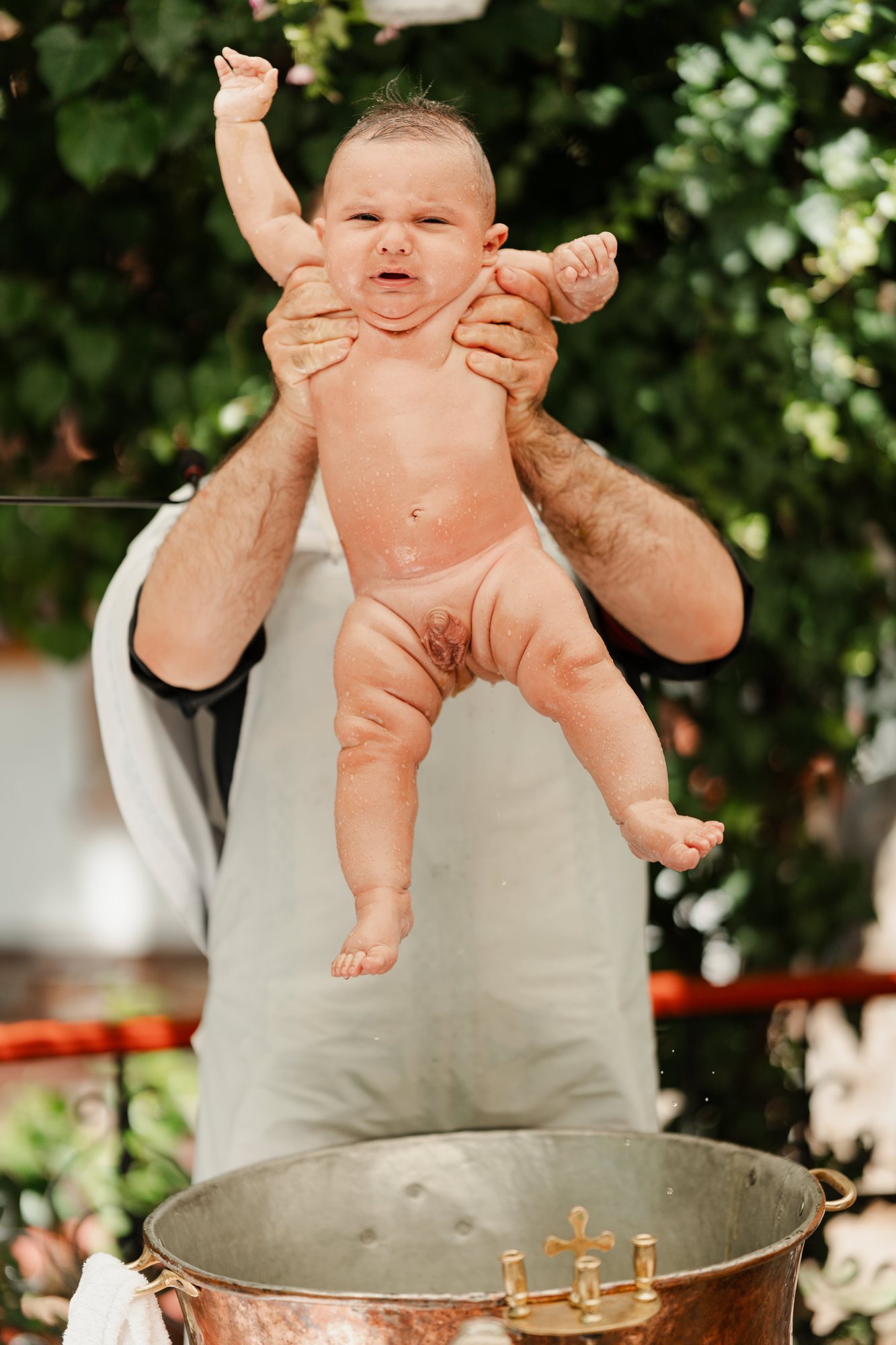 The priest lifts the baby above the copper baptismal font
