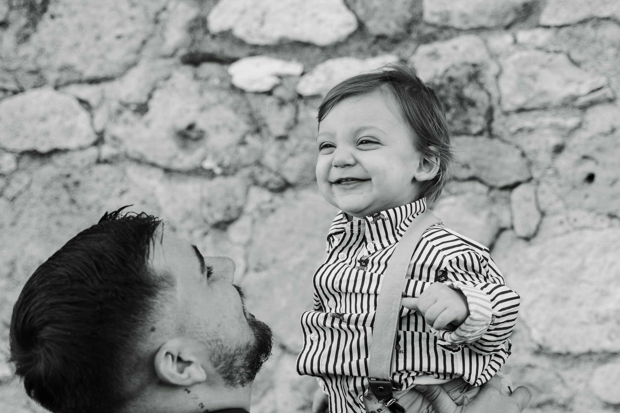 Black and white photo of a little boy laughing with his father by a stone wall
