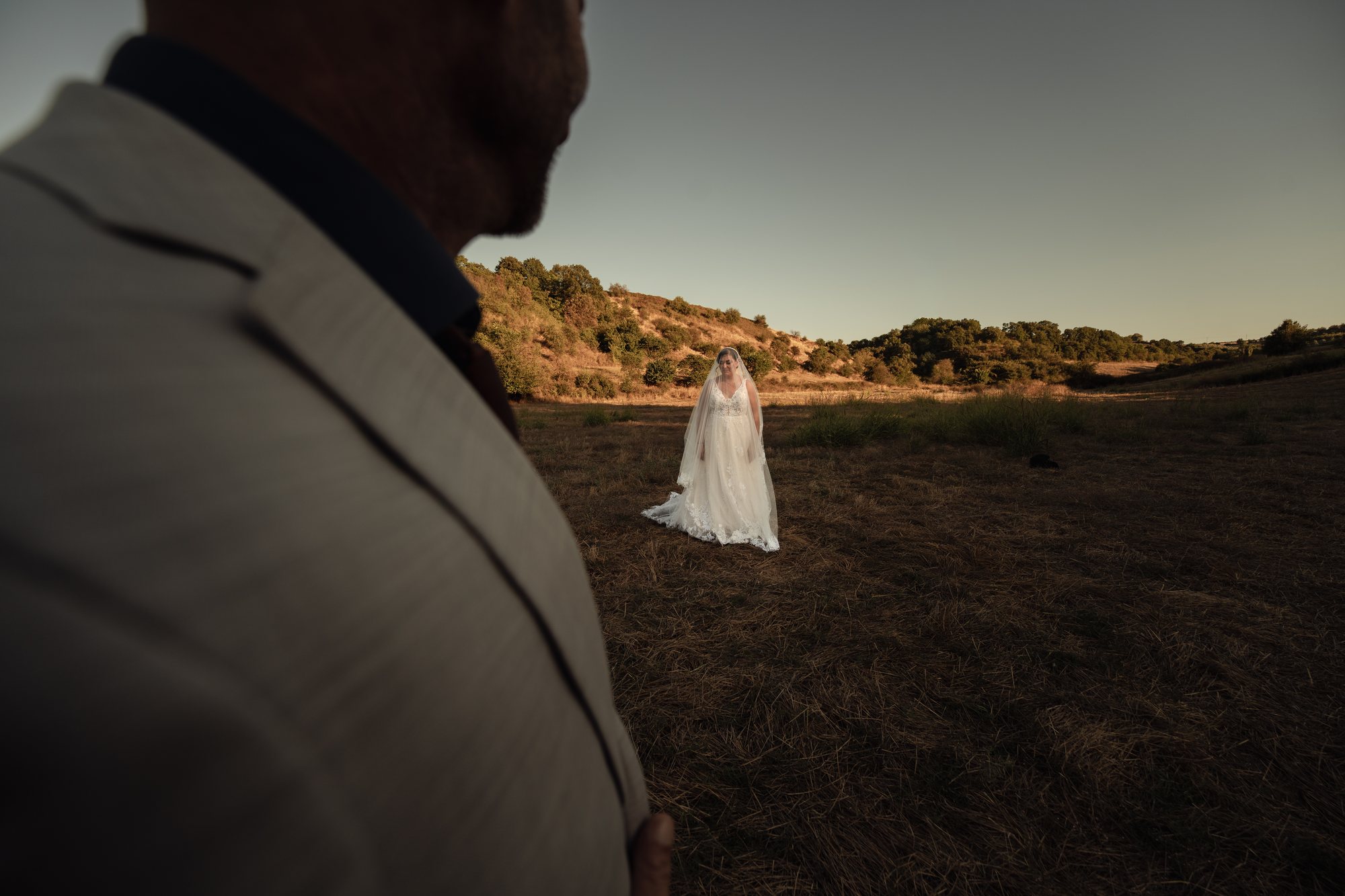The bride stands in a dry field, seen past the groom’s shoulder