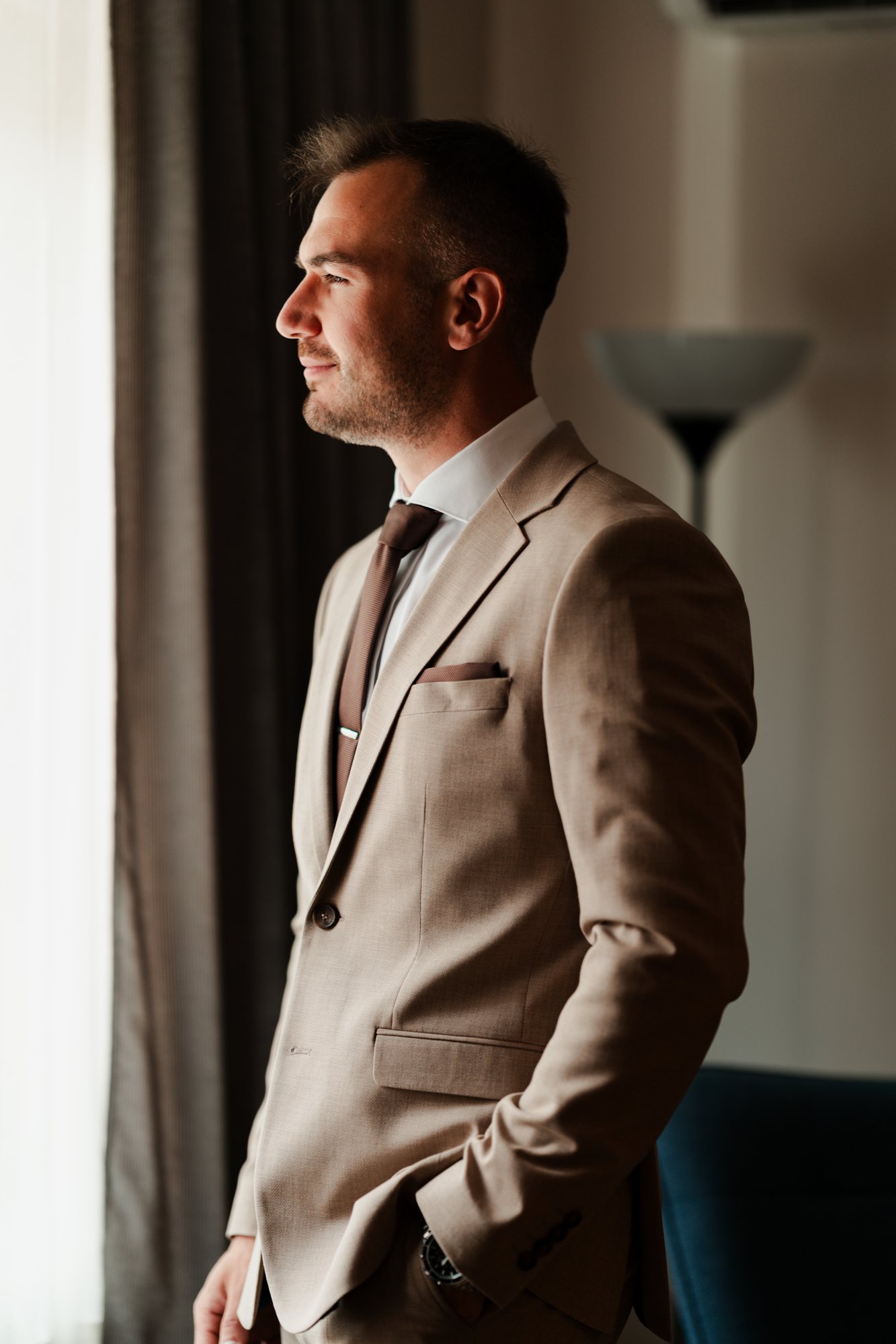The groom in a beige suit, in profile against the curtains