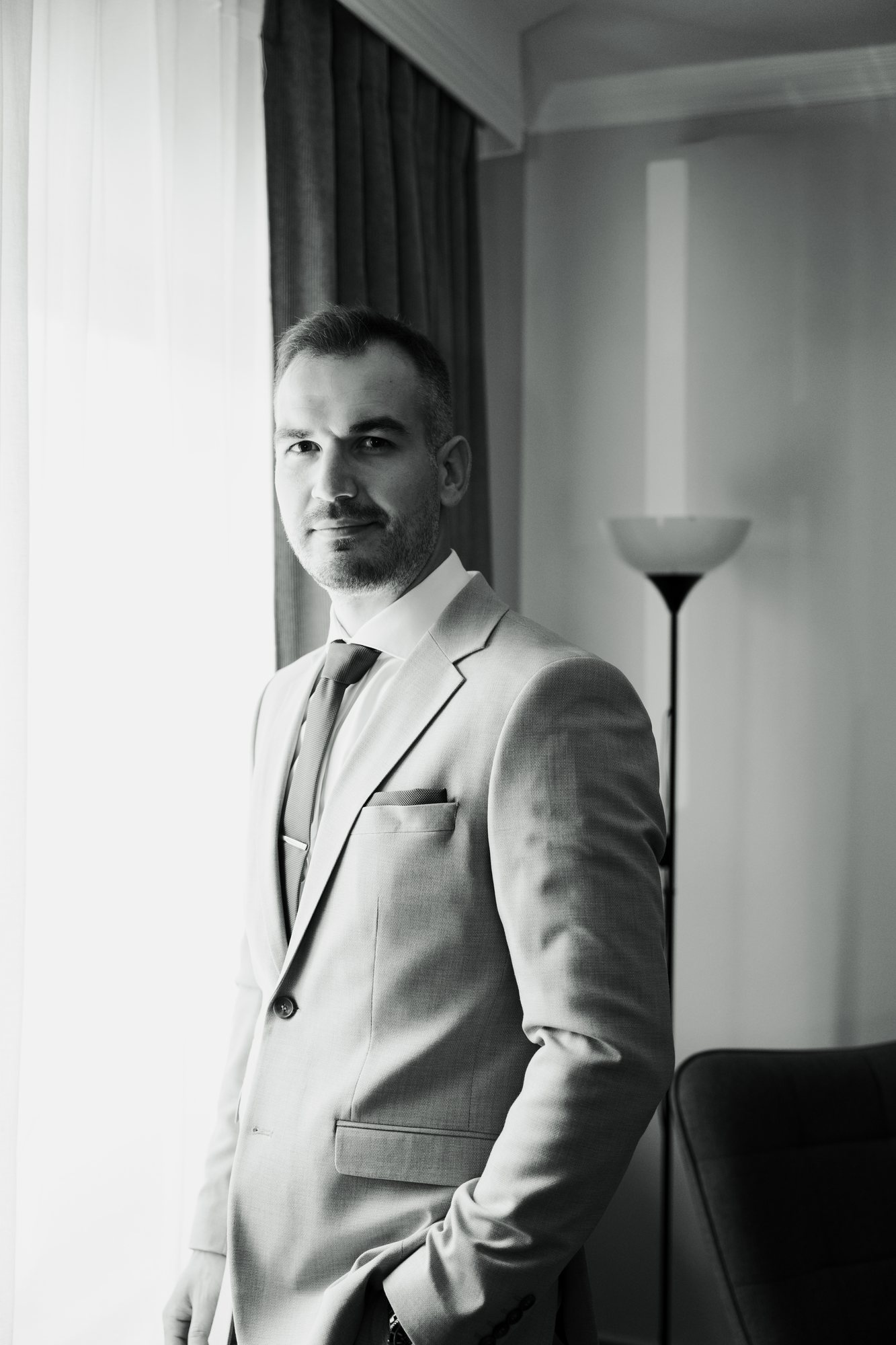 Black and white portrait of the groom standing by a bright window