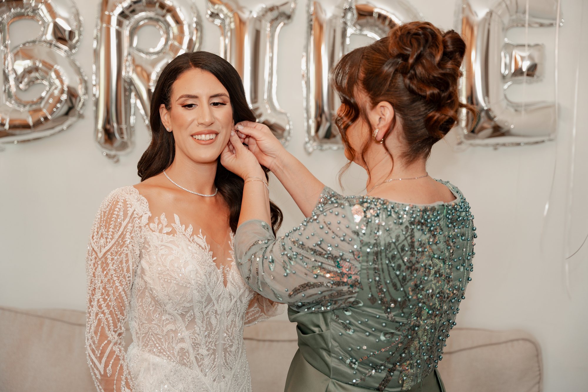 The bride’s mother fastens her earrings in front of a BRIDE balloon sign